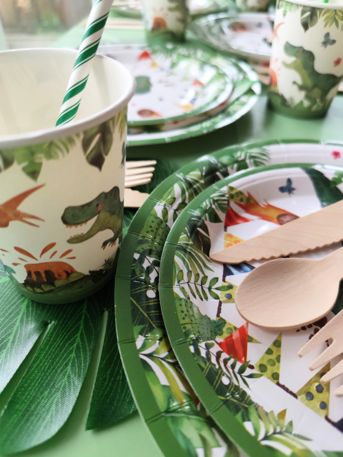 Dinosaur party themed tableware, including, plates cutlery and napkins, dinosaur cups and green striped straws. Leafy green and vibrant dinosaur patterns arranged on a table. 